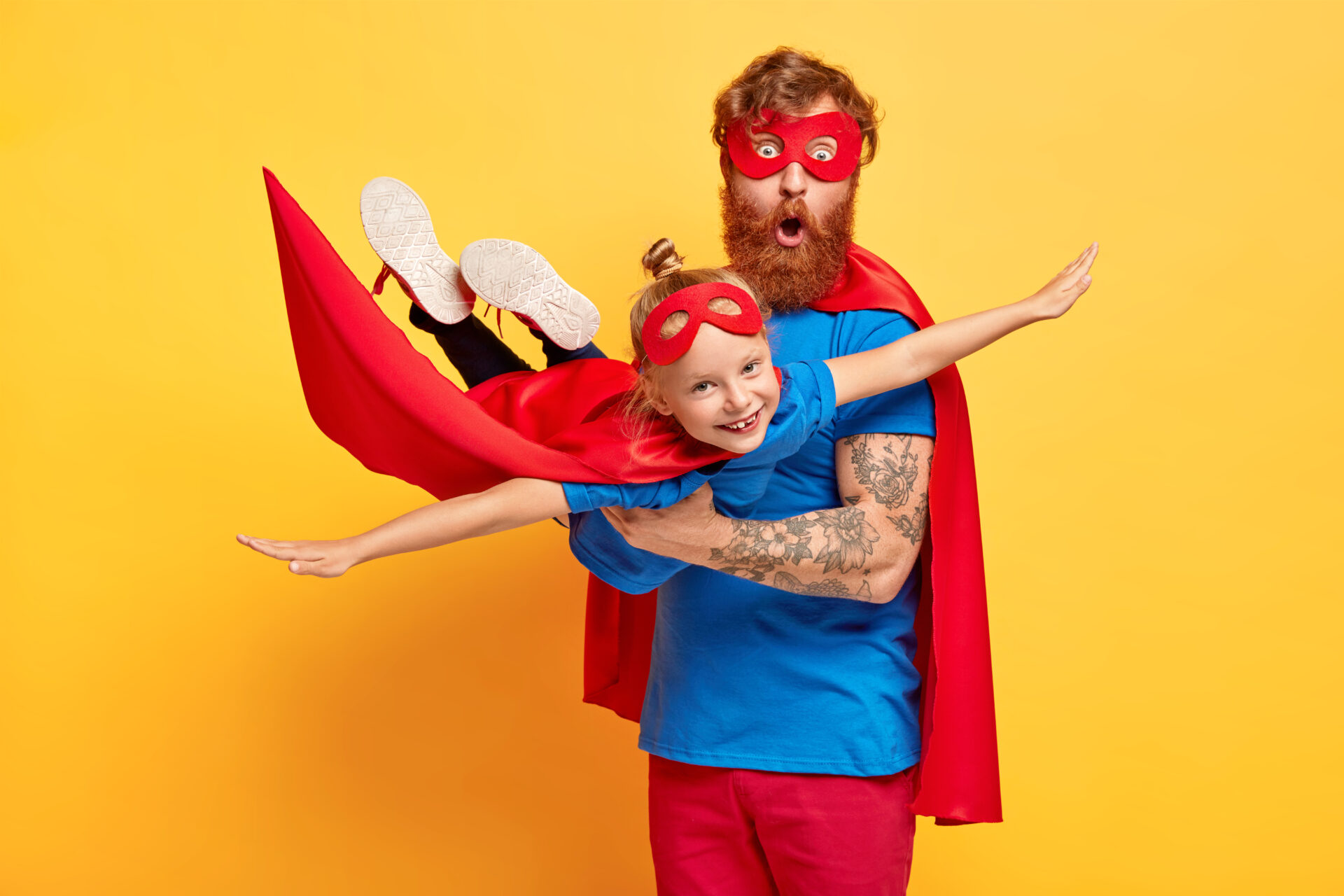 Image of ginger father and daughter dressed in superheroes, surprised man carries little kid on hands, pretend flying and saving world, pose against yellow background. Childhood and fun concept